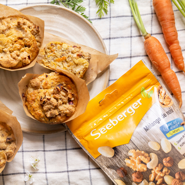 Des muffins sur une assiette reposent à côté d'un paquet de noix Seeberger, décoré de carottes sur une nappe.