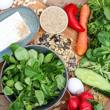 Frisches Gemüse, Samen und Nüsse liegen auf einem Holzbrett neben einer Schüssel.