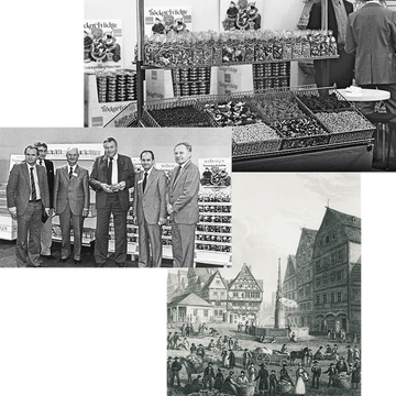 Männer stehen in Anzügen vor Trockenfrüchte-Displays; historisches Bild von Markt mit Fachwerkhäusern.