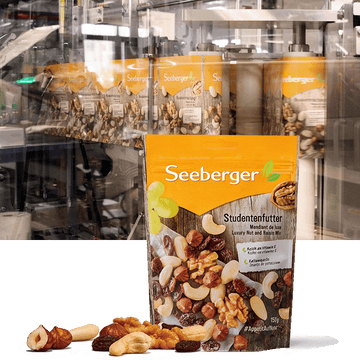 Eine Packung Seeberger Studentenfutter steht vor einer Nuss-Mischung neben Produktionsmaschinen.