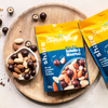 Zwei Packungen des share-Mixes liegen auf einem Holzbrett - in einer kleinen Schale danebenbefinden sich Nüsse und schokolierte Haselnusskerne